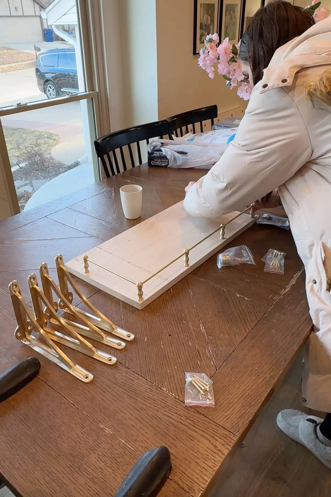 The woman continues working on building a brass rail shelf, with gold brackets and other supplies laid out on the dining table. A cup of coffee sits nearby